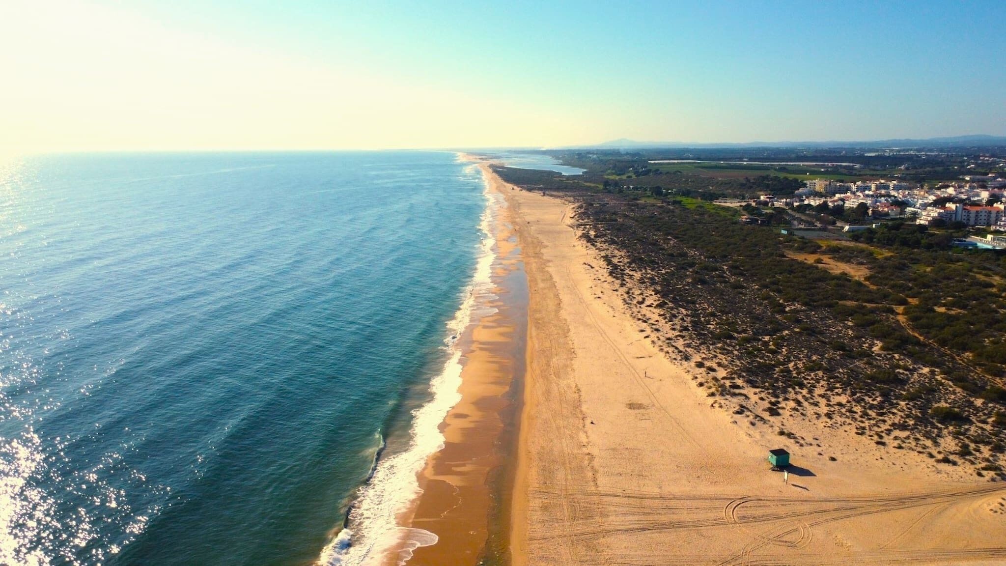 Luchtbeeld van een strand met een lange zandvlakte, enkele verspreide gebouwen en een helder uitzicht op de oceaan.