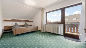 Une pièce avec un plafond en pente, un lit en bois avec des oreillers, un radiateur, une grande fenêtre donnant sur des immeubles et un tapis à motifs verts.