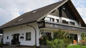Une maison à deux étages, dotée d’un toit en pente, d’un balcon avec une balustrade et d’un porche à l’avant avec des marches.