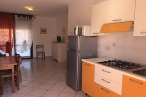 Une cuisine compacte équipée d’un réfrigérateur, d’un four à micro-ondes et d’une cuisinière à gaz. On y trouve une table à manger avec des chaises et une fenêtre avec des rideaux.