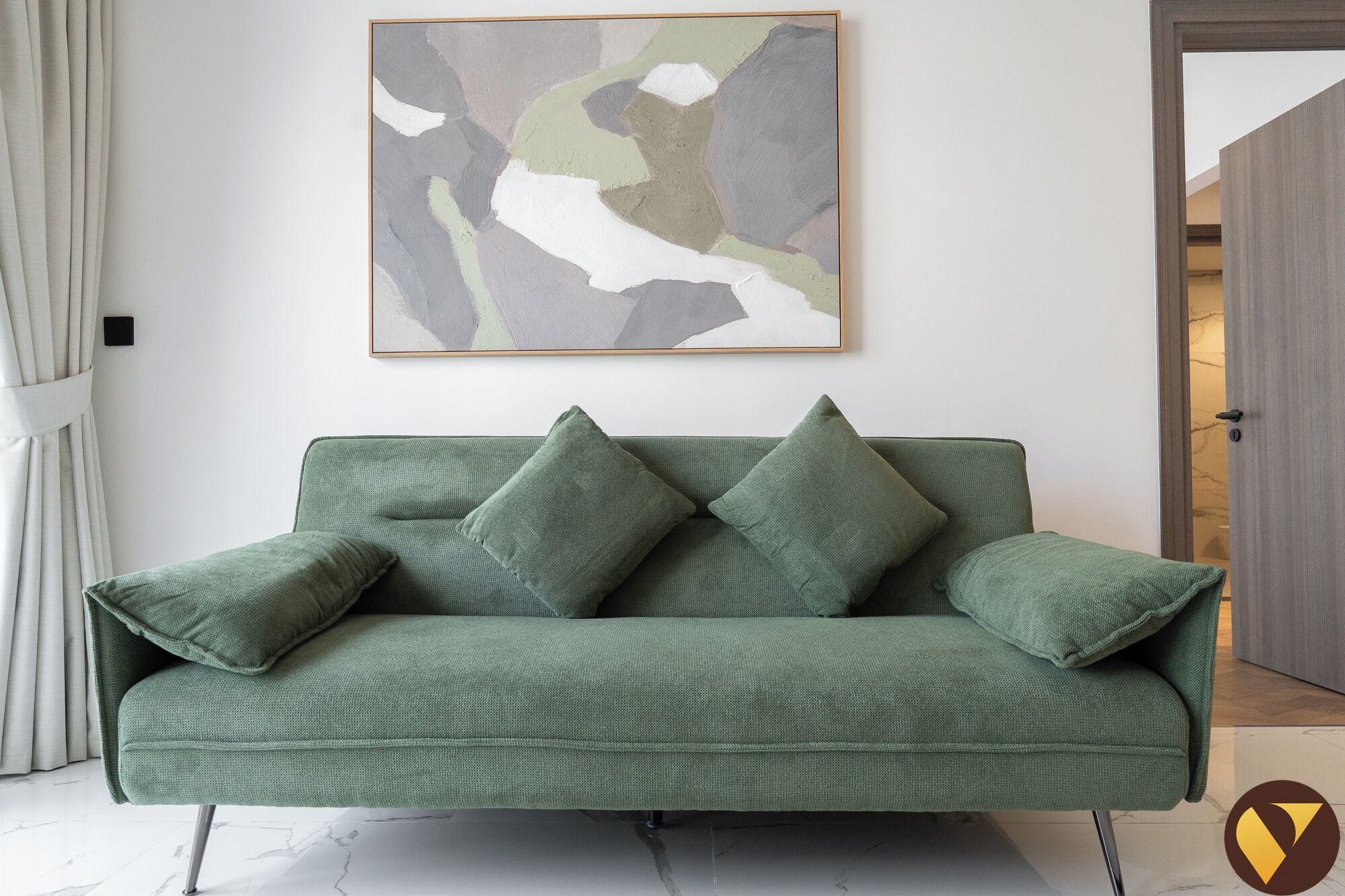 Un canapé vert avec quatre coussins, un tableau abstrait encadré au-dessus, et une porte sur la droite.