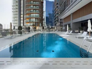 A modern indoor swimming pool with lounge chairs and umbrellas, surrounded by high-rise buildings. - Vogue | Royal Regency Business Bay near Downtown (Dubai)