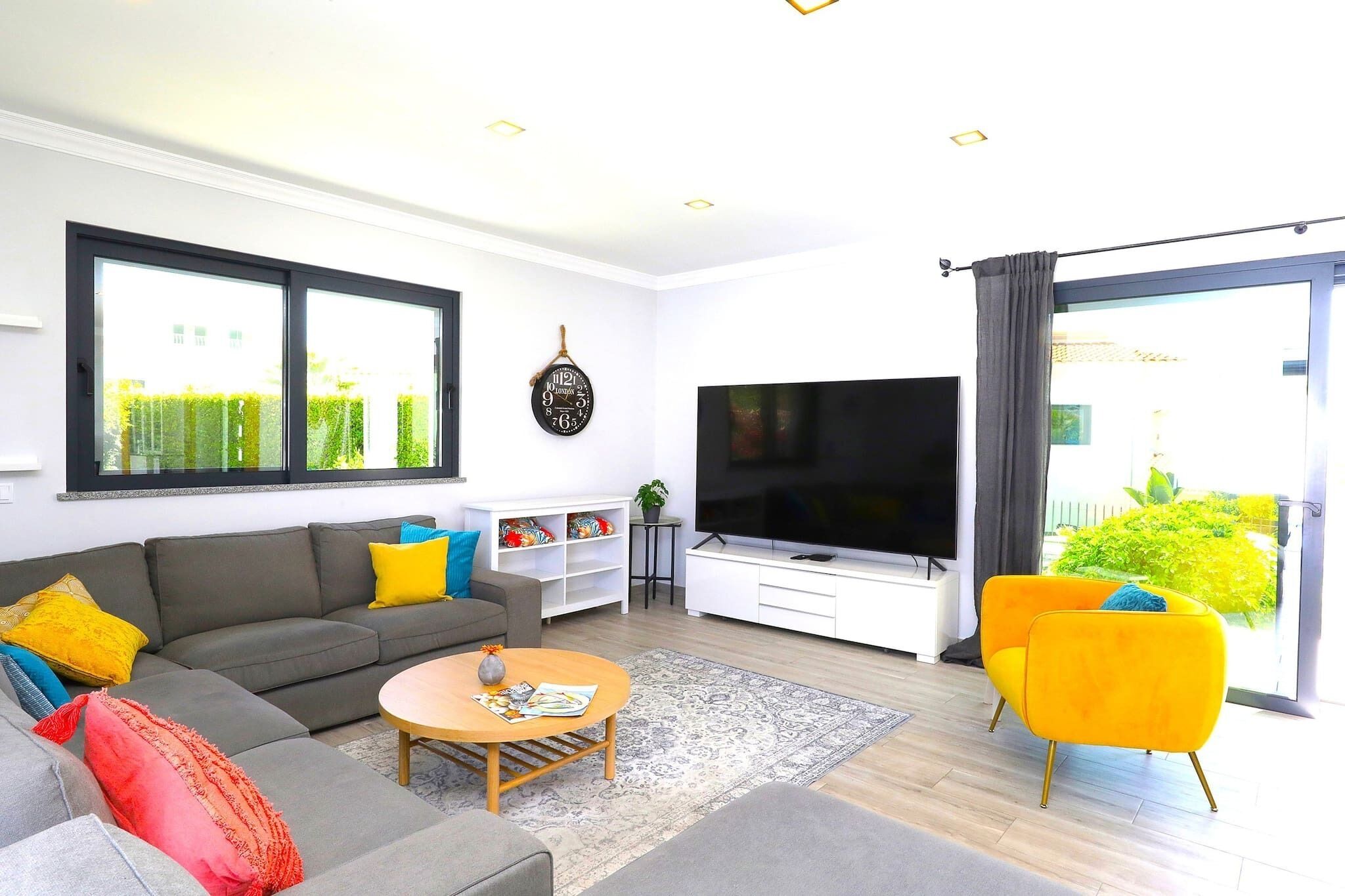 A modern living room with a grey sofa, yellow armchairs, a flat-screen TV, and a large window.