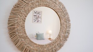 Un espejo redondo con marco tejido, una cama con almohada azul, una lámpara de noche y un cuadro enmarcado en la pared.