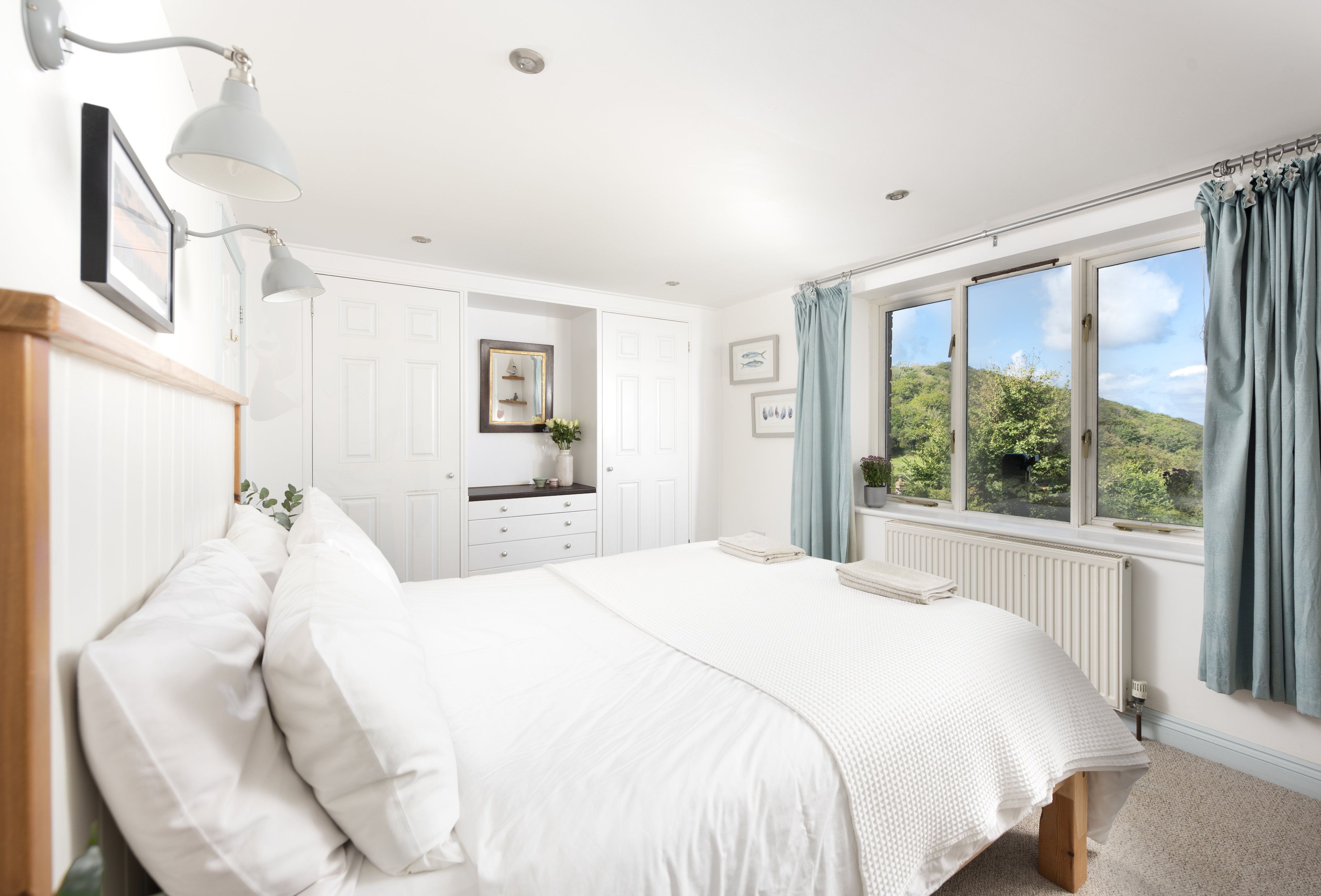 Une chambre à coucher avec un lit, une commode, une fenêtre avec des rideaux et une vue sur la verdure à l’extérieur.