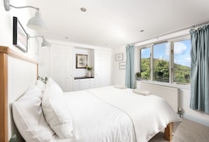 Ein Schlafzimmer mit Bett, Kommode, einem Fenster mit Vorhängen und Blick ins Grüne.