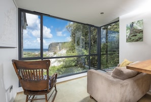 A room with a large window offering a view of the sea and cliffs. There is a wooden chair and a sofa with cushions. - Escape to coastal bliss at this stylish family retreat of Lower Hillcott (Bude)