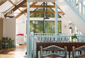 A spacious room with a high ceiling, wooden beams, a staircase, and a large window offering a view of greenery. - Escape to coastal bliss at this stylish family retreat of Lower Hillcott (Bude)
