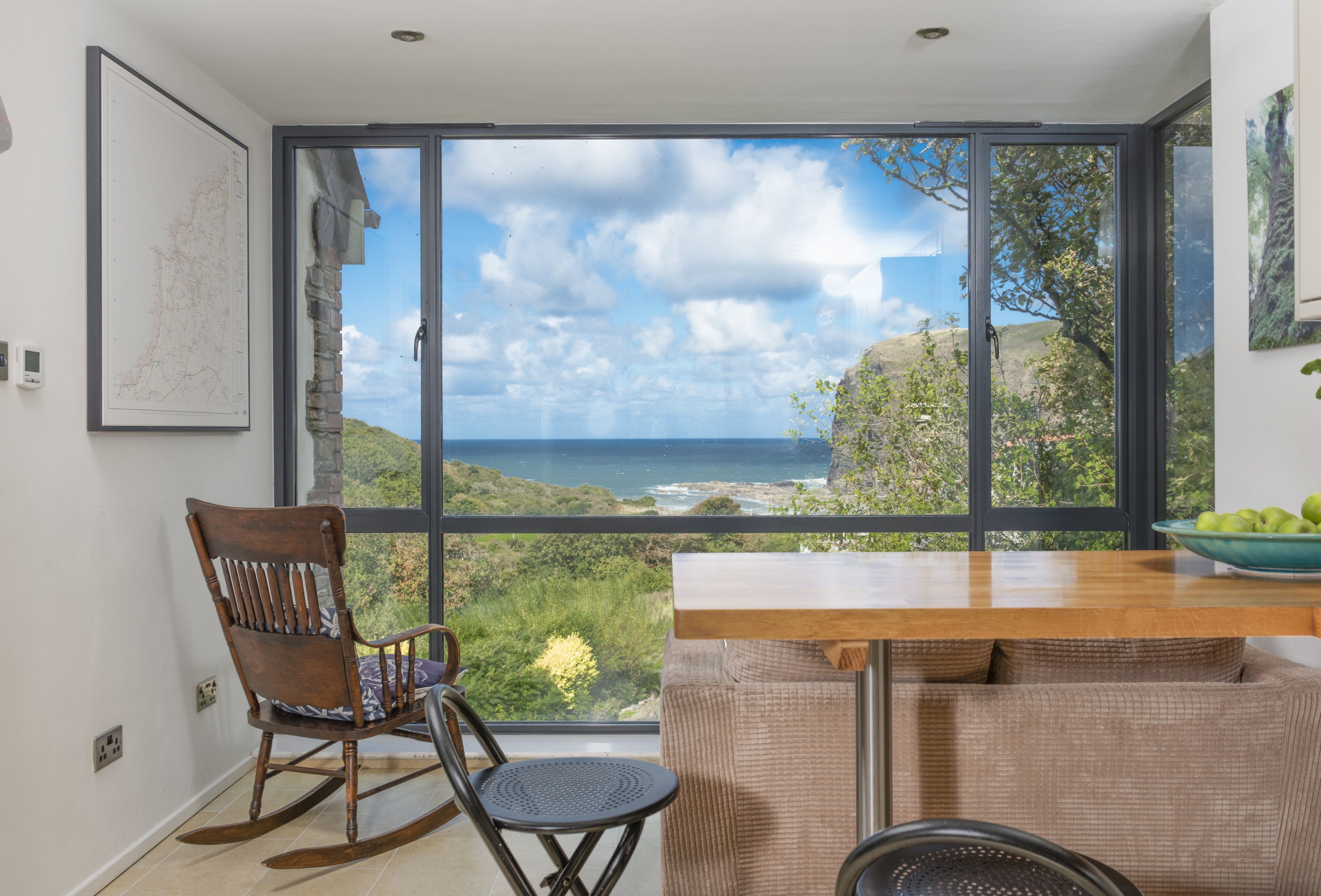 A room with a large window overlooking a coastal landscape, a wooden rocking chair, a dining table, and a bowl of fruit.