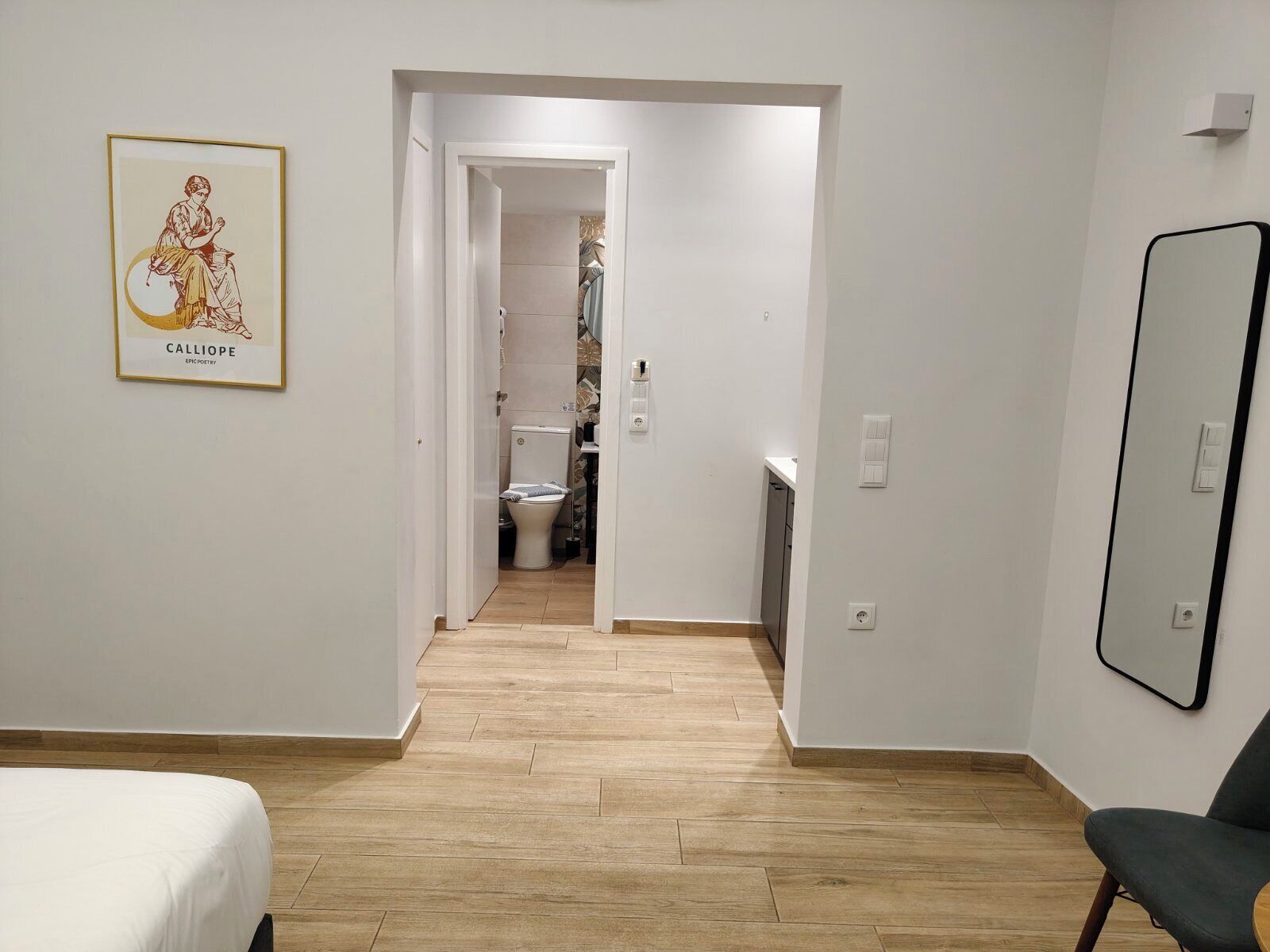 A modern hotel room with a wooden floor, a bed, a chair, a bathroom with a toilet and sink, and a framed picture on the wall.