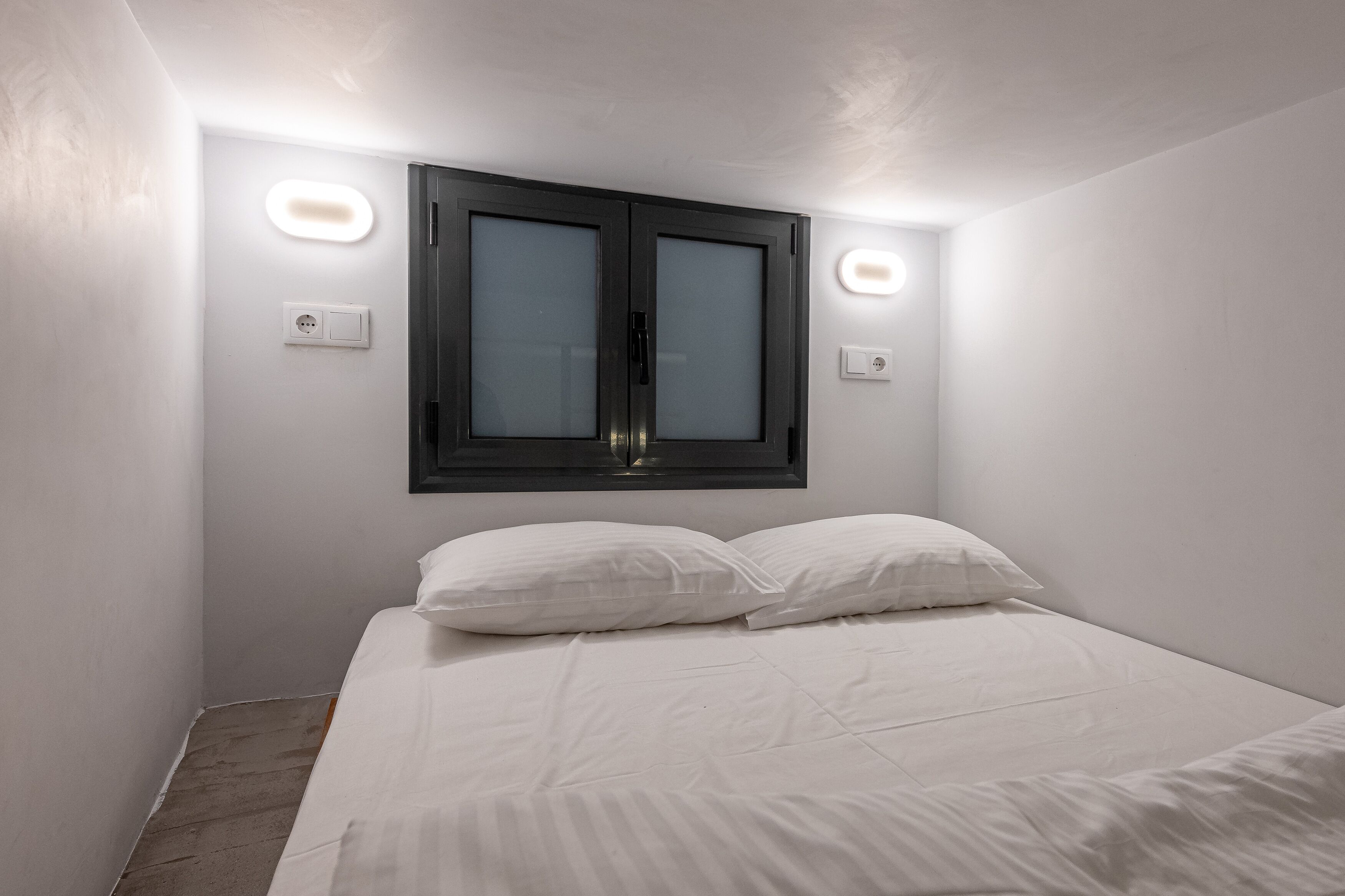 A bedroom with a bed, white walls, a window with frosted glass, and two wall-mounted lights.