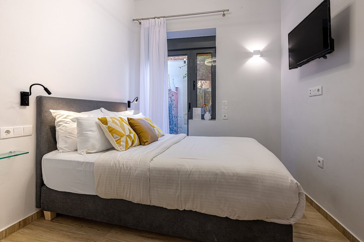 A neatly made bed with a grey headboard, white and yellow pillows, and a white duvet. A flat-screen TV is mounted on the wall to the right.