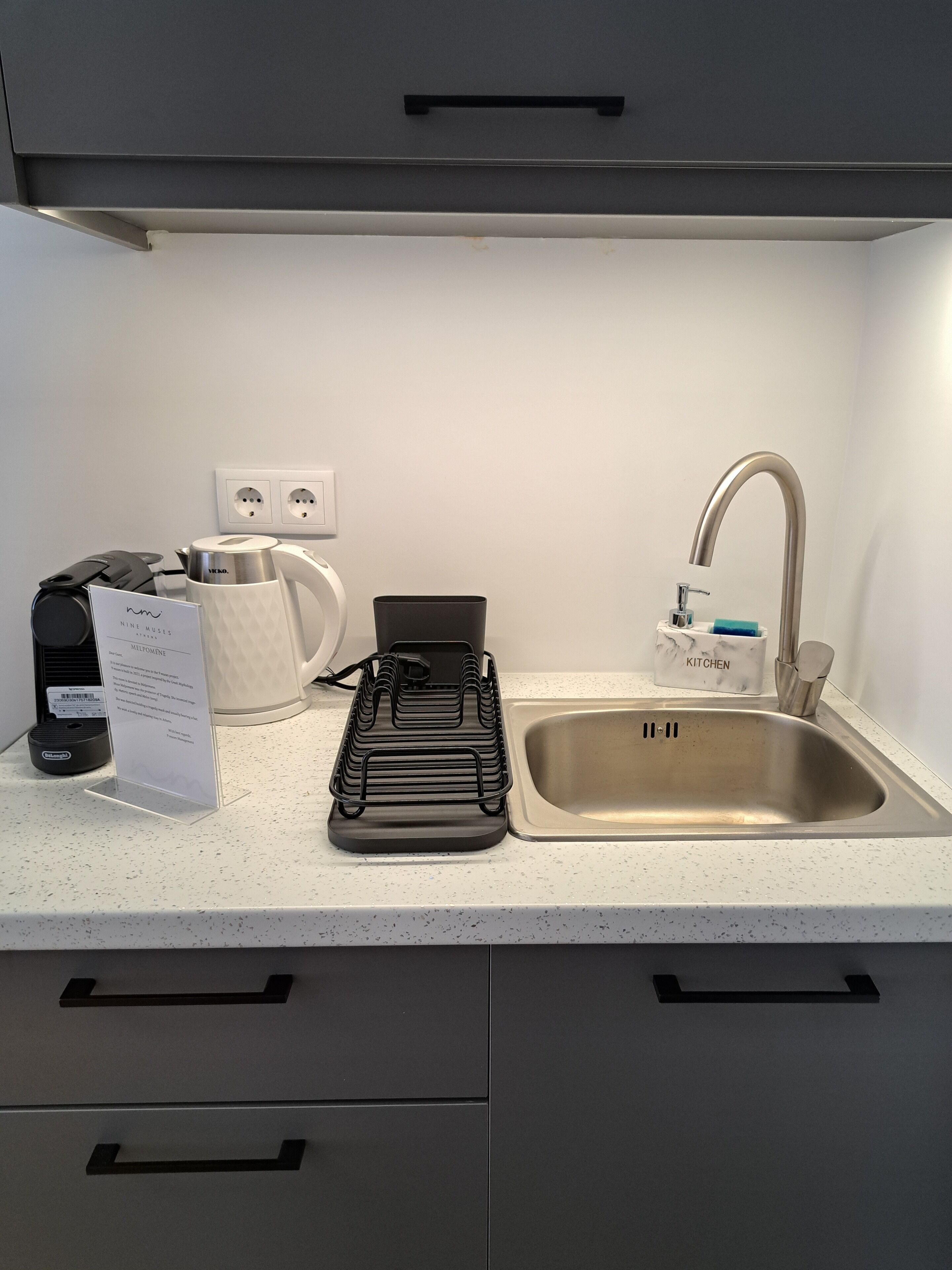 Una cocina con un lavamanos de acero inoxidable, una tetera, una cafetera y una bandeja negra. Hay dos cajones debajo de la encimera.