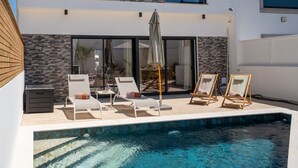 Un espace piscine extérieur moderne, doté de chaises longues et de parasols, d’un mur en pierre et d’une grande fenêtre donnant sur la piscine.