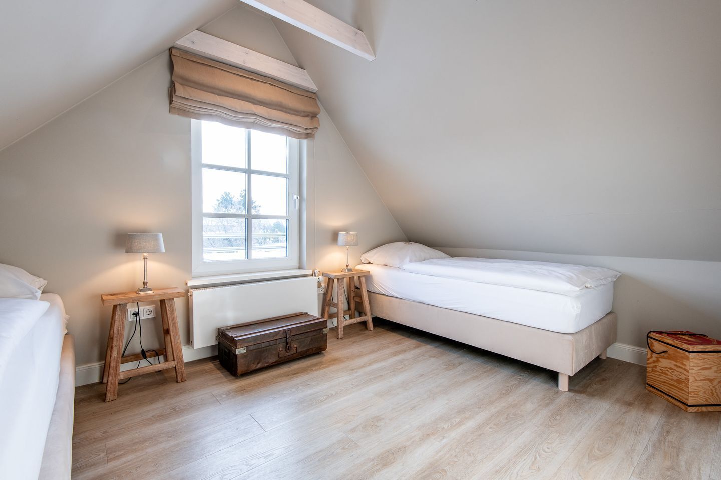 A small, cozy attic bedroom with a sloped ceiling, a window with a beige valance, a wooden bench, a small table with a lamp, and a suitcase.