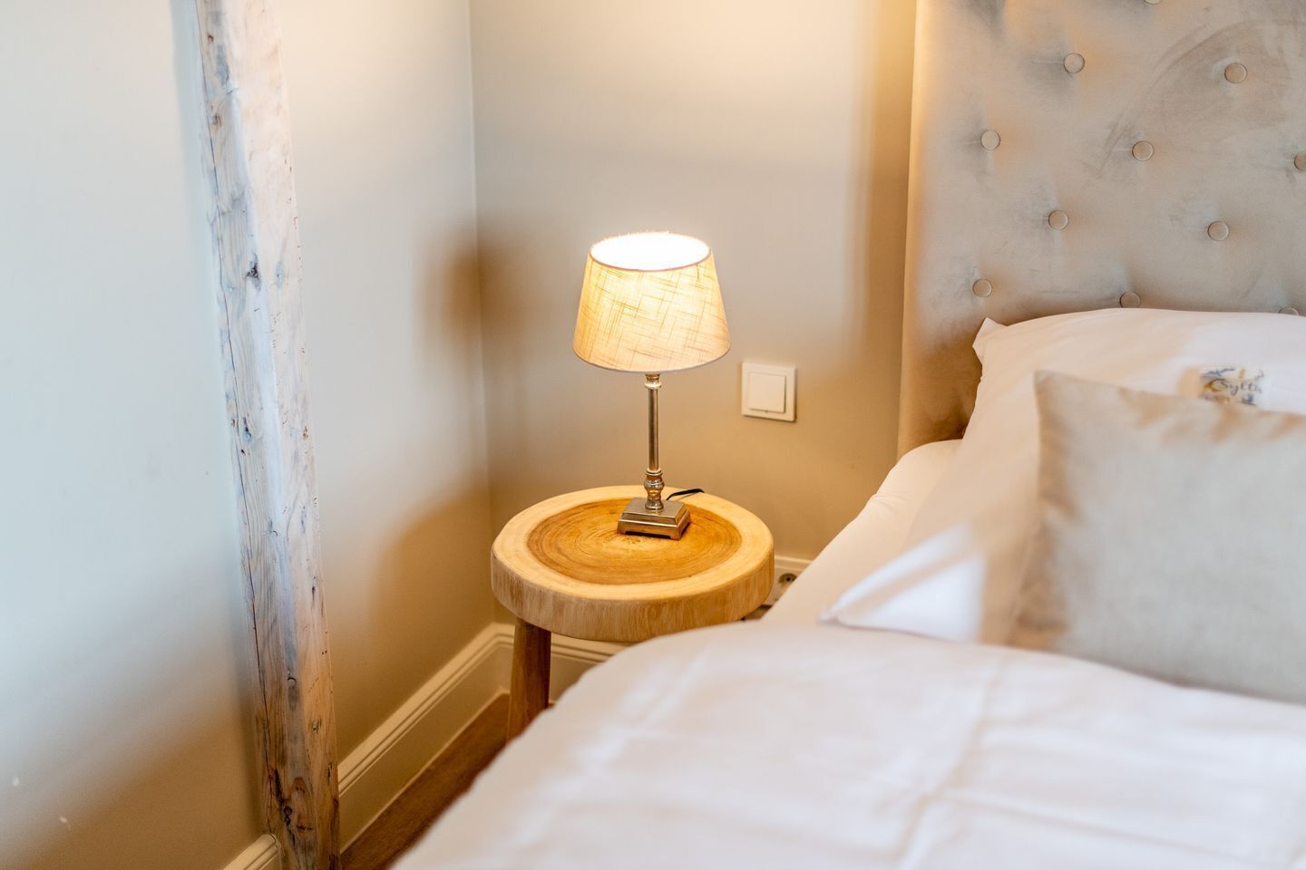 A neatly made bed with white linens, a round wooden bedside table with a lamp, and a tufted headboard.