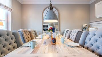A dining area with a long wooden table, upholstered chairs, a hanging light fixture, and a kitchen in the background.