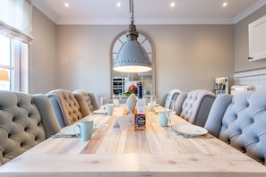 A dining area with a long wooden table, upholstered chairs, a hanging light fixture, and a kitchen in the background. - House SyltZauber, Germany (Sylt)