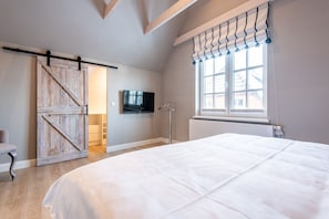 A bedroom with a large bed, a sliding barn door closet, a TV mounted on the wall, and a window with curtains. - House SyltZauber, Germany (Sylt)