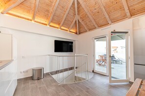 Un intérieur moderne avec un plafond en bois, un téléviseur à écran plat fixé au mur, une poubelle et une porte-fenêtre coulissante donnant sur un espace extérieur équipé d’une table et de chaises.