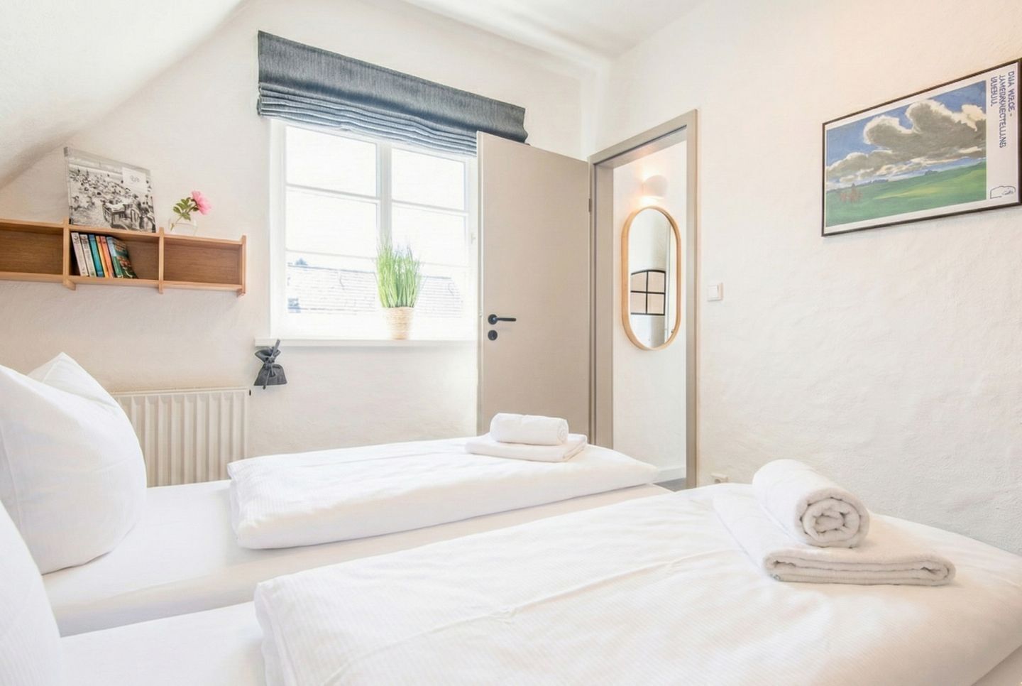 A hotel room with two beds, a window with a view, a bookshelf, a radiator, and a mirror.