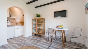 Una cocina moderna con armarios blancos, un mueble de madera con puertas de cristal, una mesa redonda con un frutero y un televisor montado en la pared.