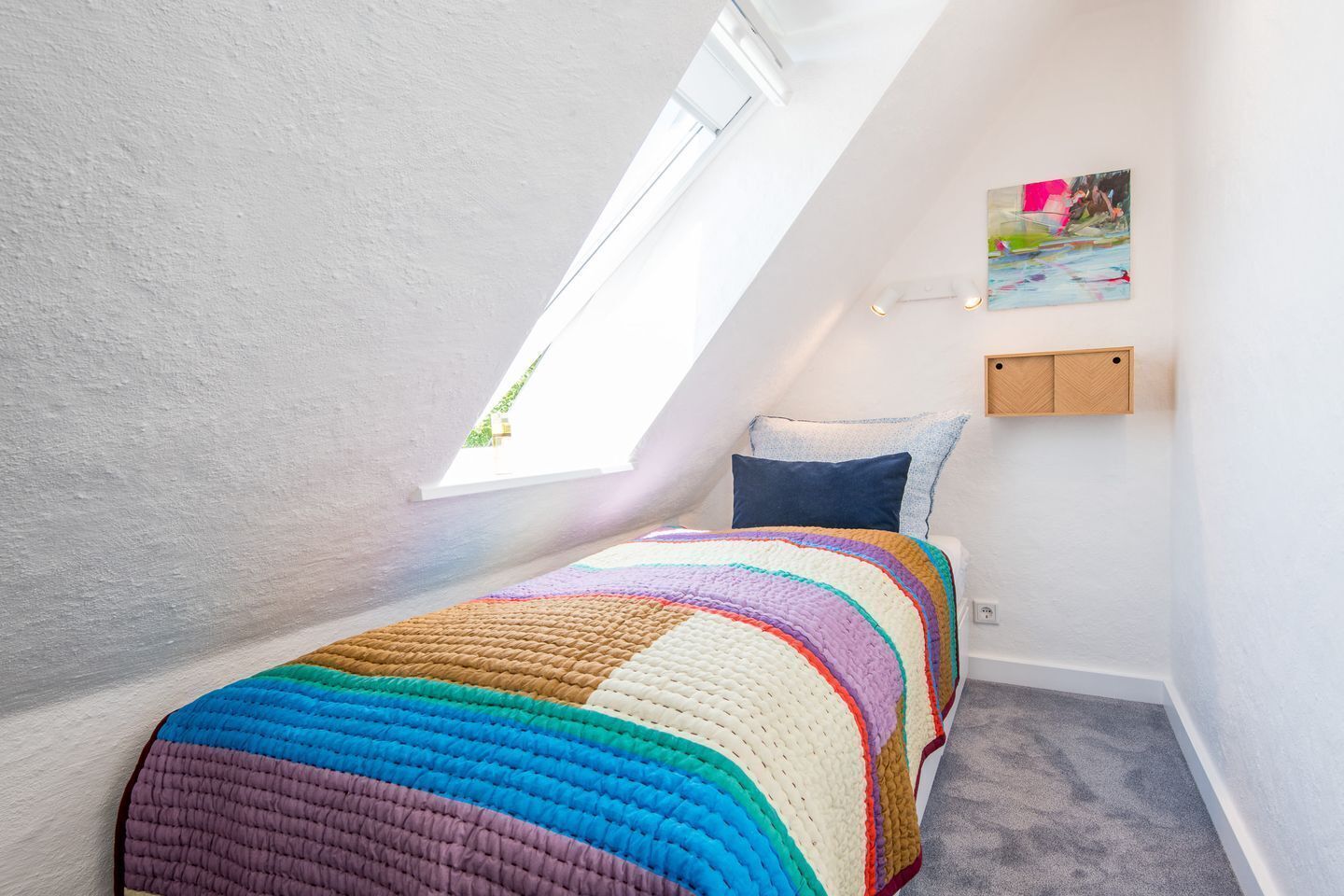 A small, bright room with a sloped ceiling and a skylight, featuring a colorful quilted bedspread, a single pillow, and a small wooden shelf with a painting.