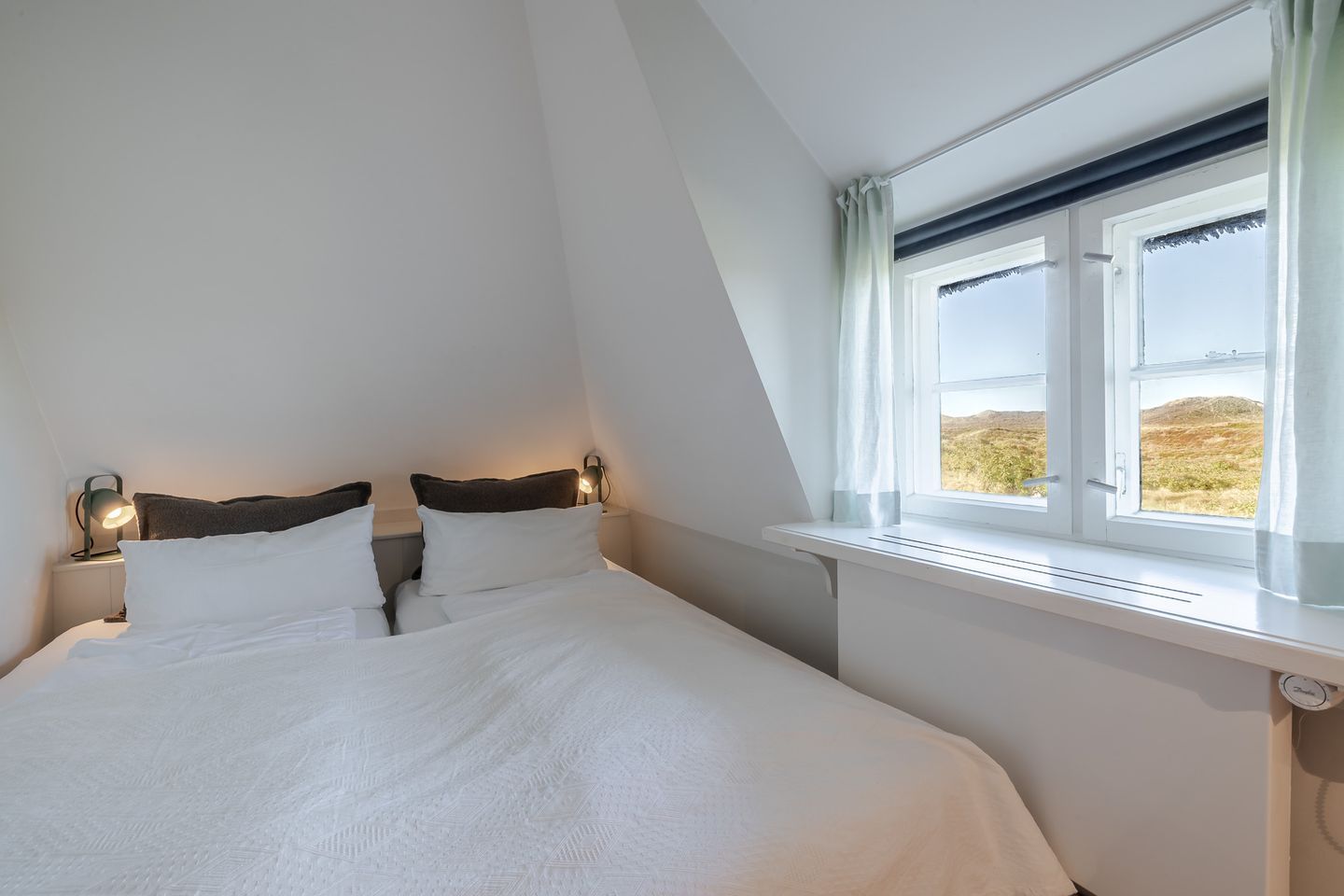 A bedroom with a bed, two bedside lamps, a window with a view of the countryside, and a white ceiling.