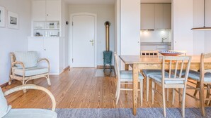 A cozy living room with a wooden dining table, chairs, and a sofa. A kitchen area is visible in the background with white cabinets and a stove. - Kleiner Dünenhof, Germany (Kampen (Sylt))