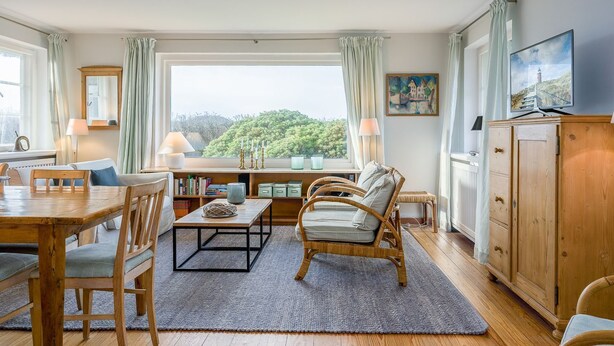 A cozy living room with a wooden dining table, comfortable seating, a television, and a view of greenery outside. - Kleiner Dünenhof, Germany (Kampen (Sylt))