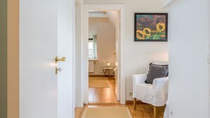 A hallway with a white door, a framed painting of sunflowers, and a white chair with a cushion. - Kleiner Dünenhof, Germany (Kampen (Sylt))