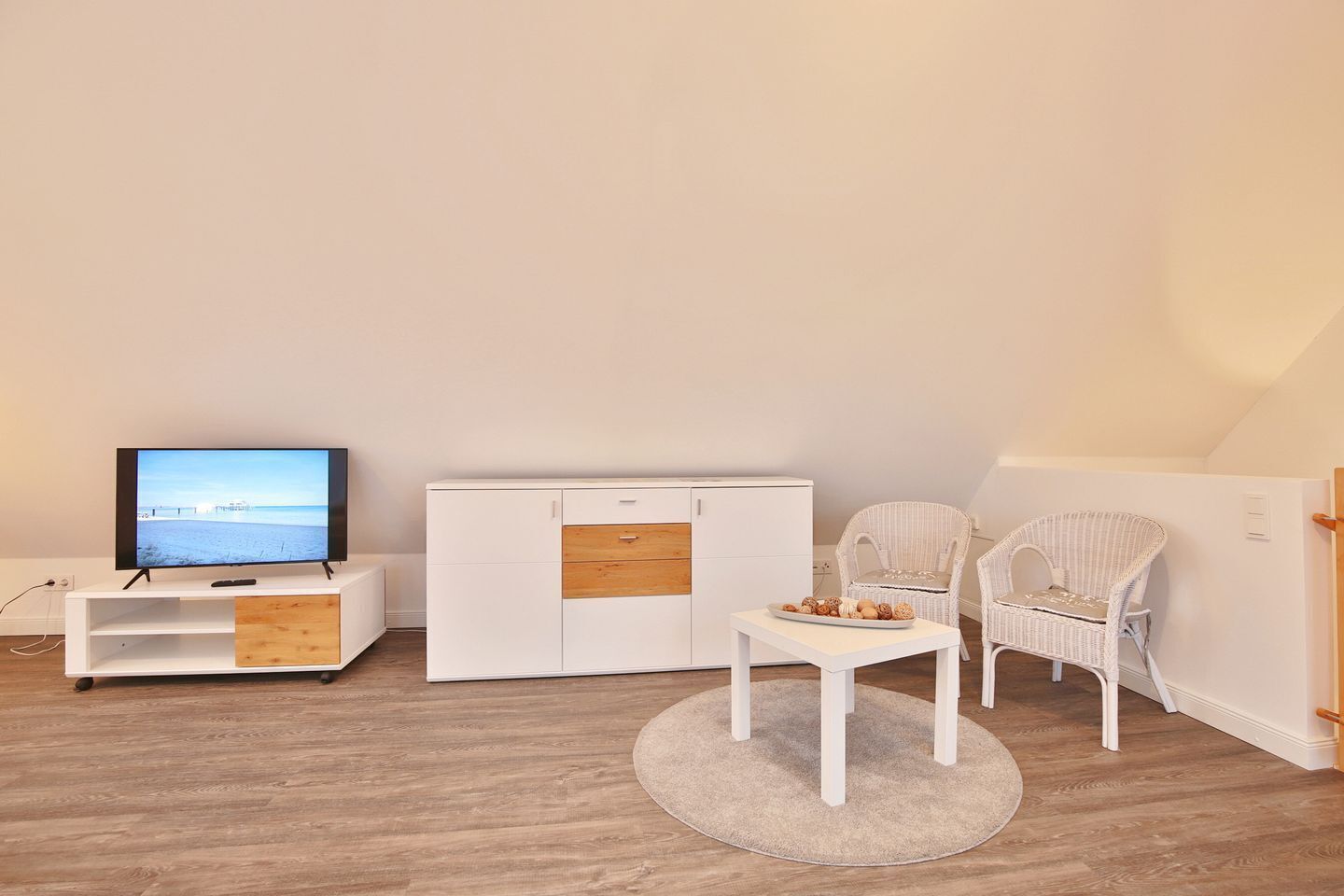 A room with a TV on a stand, a white wardrobe, two wicker chairs, and a small table with a plate of pastries.