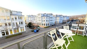 Un balcón con césped artificial y sillas blancas, con vistas a una calle y edificios de estilo europeo tradicional.