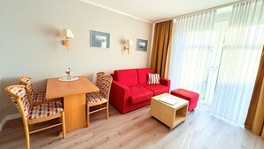 Una habitación con una mesa de comedor de madera, dos sillas, un sofá rojo, un pequeño otoman y una ventana con cortinas traslúcidas.