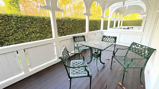 Eine überdachte Veranda mit Tisch und Stühlen, umgeben von weißen Geländern und einer Hecke.