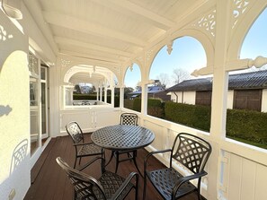 Eine überdachte Terrasse mit rundem Tisch und vier Stühlen, eine weiße Außenwand und Blick auf benachbarte Häuser.