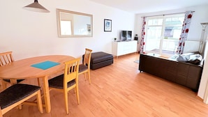 A living room with a wooden dining table, two chairs, a sofa, a TV, and a window with curtains. - Strandresidenz Juliusruh apartment 11- ground floor, balcony, 2 bedrooms - family friendly (Breege)