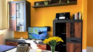A modern living room with a TV, a wooden cabinet, a bookshelf, and a vase with yellow flowers. - Fewo Ostseelaune - directly by the forest and the sea - Ostseelaune - Comfortable apartment (Dranske)