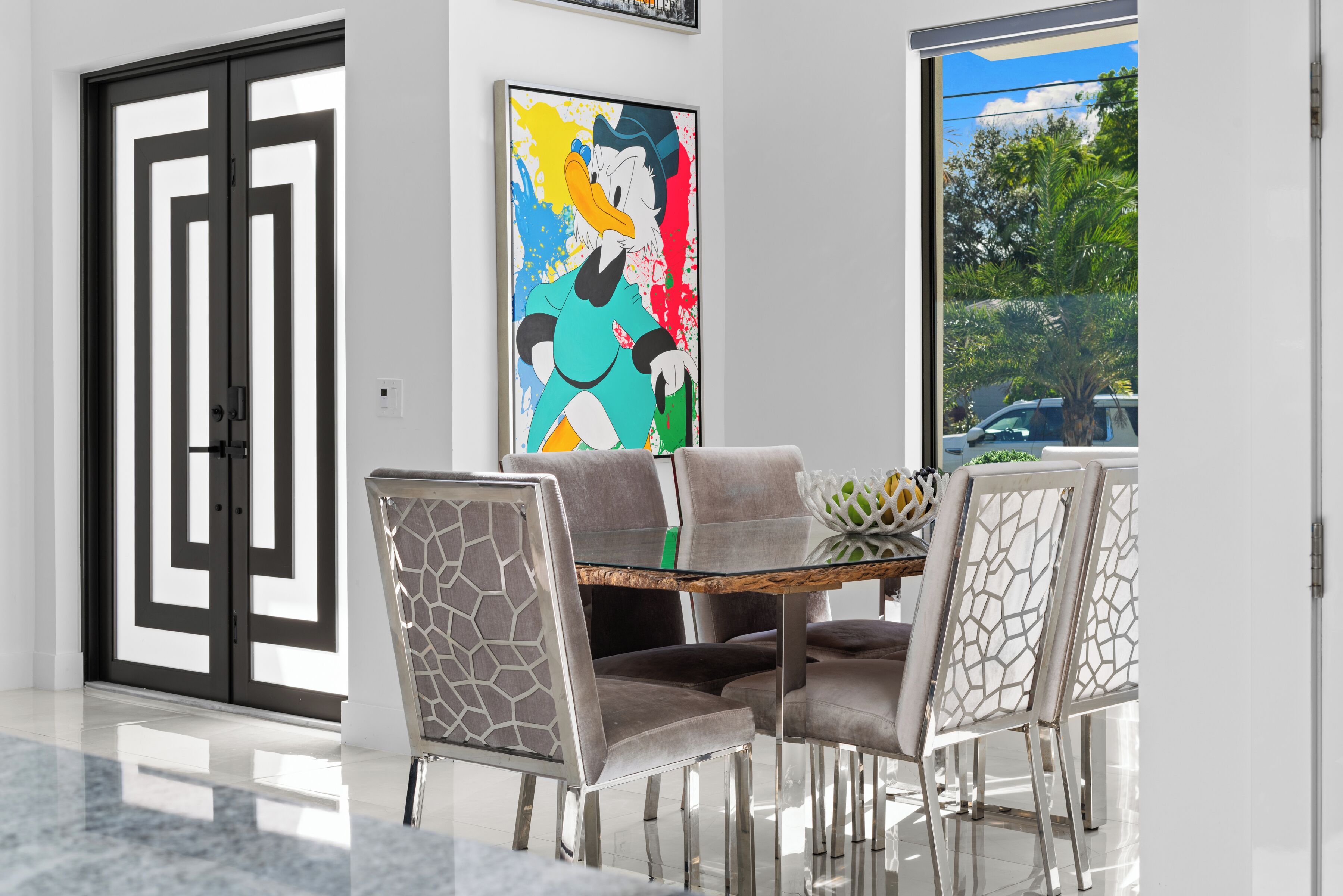 Un coin repas avec une table à plateau en verre, quatre chaises rembourrées ornées de motifs géom étriques, une peinture abstraite aux couleurs vives accrochée au mur et une vue sur l’extérieur grâce à une grande fenêtre.