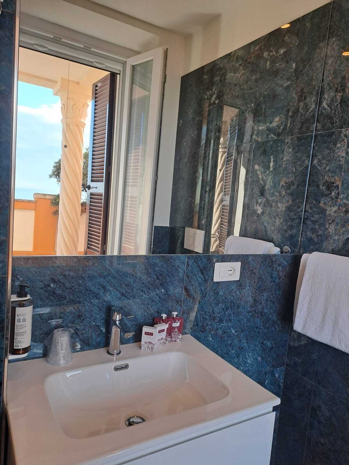 Un baño con un lavamanos blanco, paredes de mármol oscuro, un espejo y una ventana con persianas.