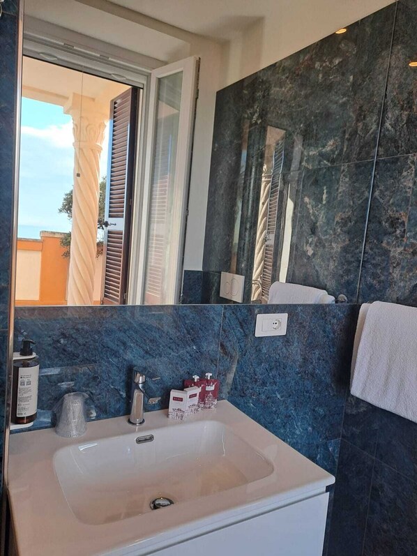 A bathroom with a white sink, dark marble walls, a mirror, and a window with blinds. - Delightful beachfront apartment equipped with all the comforts (Lido di Ostia)