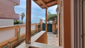 A covered outdoor area with a wooden ceiling, a concrete bench, and a view of a building with a green gate and palm trees. - Delightful beachfront apartment equipped with all the comforts (Lido di Ostia)