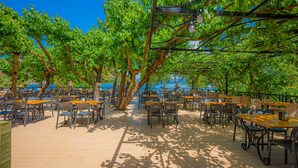 An outdoor café with tables and chairs under trees, overlooking a body of water. - Golmar Beach& Spa Hotel (Marmaris)