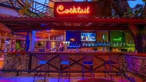 An outdoor bar area with a neon sign reading "Cocktail," a bar counter with various bottles, and several chairs.