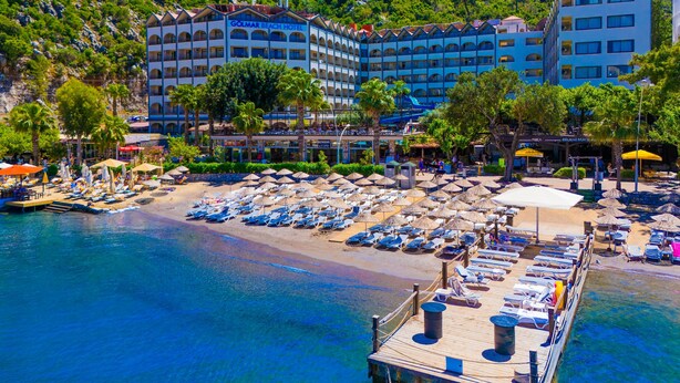 Un hotel con varios balcones con vistas a una playa, una zona de piscina con tumbonas y un muelle que se adentra en el agua.