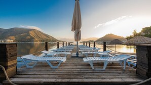 Un muelle de madera con hileras de sillas reclinables blancas y una sombrilla, con vistas a un lago tranquilo y montañas al fondo.
