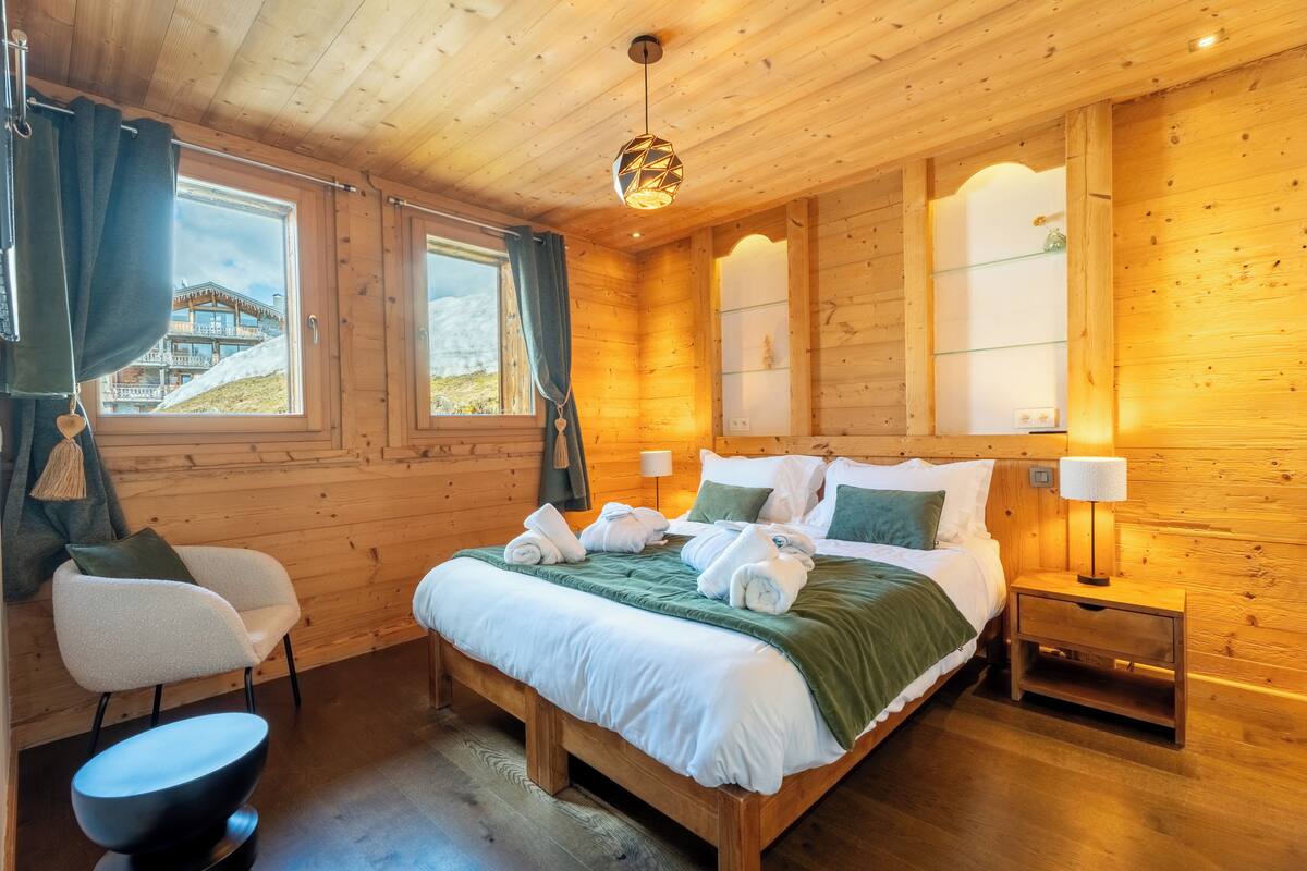 A wooden cabin bedroom with a bed, bedside tables, and a view of a building outside.