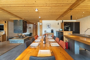 A modern dining area with a long wooden table set for a meal, a kitchen with a refrigerator and stove, and a cozy seating area with chairs and a sofa. - Hameau des clos Virolet (Les Gets)