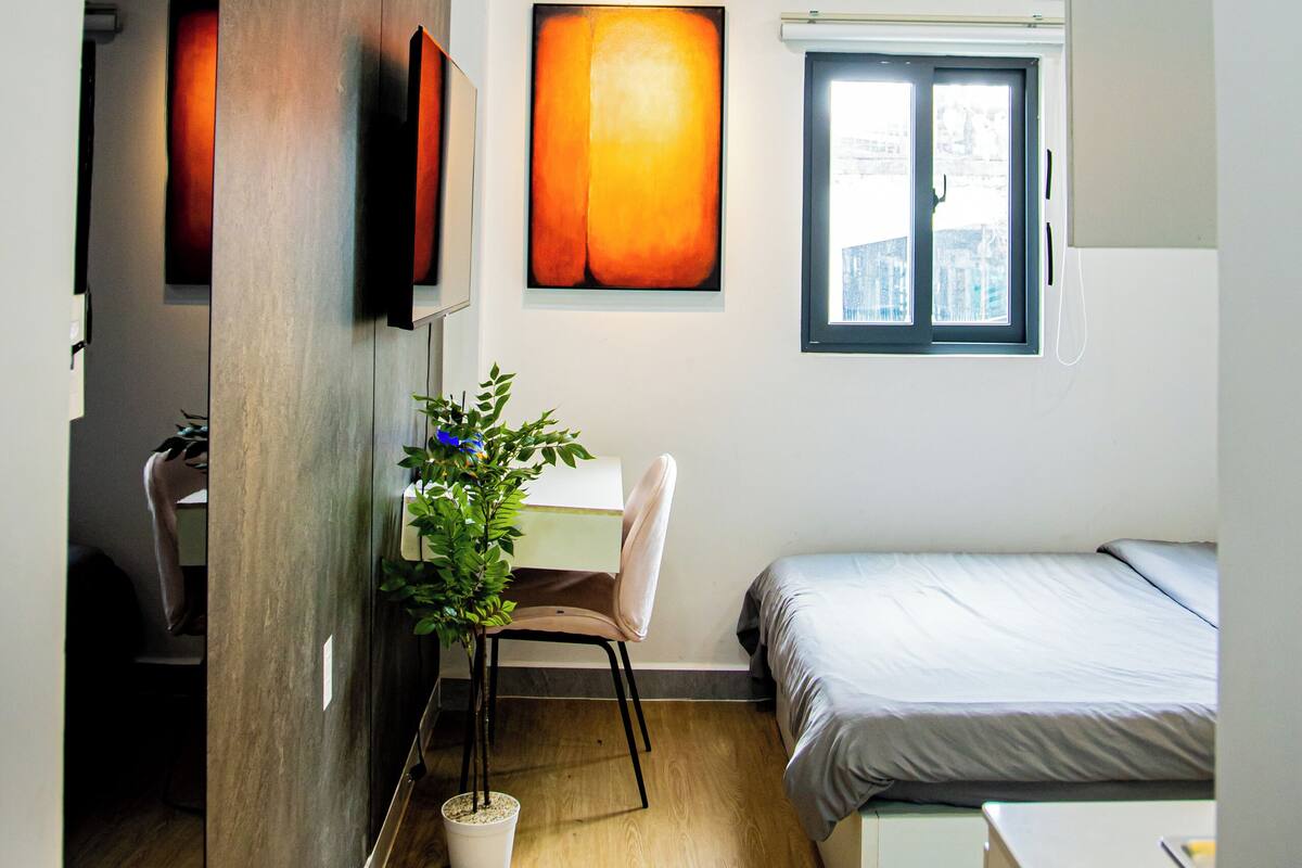 A modern bedroom with a bed, a desk, a chair, a potted plant, and a window.