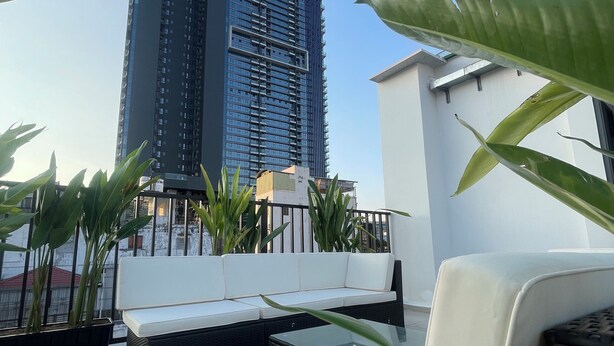 Een terras op het dak met een witte bank, een glazen tafel en potplanten. Op de achtergrond een hoog gebouw.